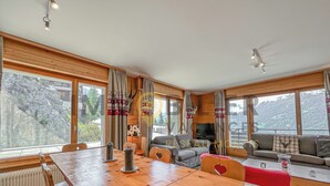 L’intérieur chaleureux d’un chalet comprend une table à manger en bois, un canapé et un téléviseur. De grandes fenêtres offrent une vue sur un paysage rocheux.