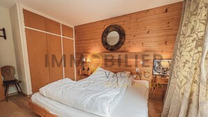 A bedroom with a wooden floor, a bed with white bedding, a wooden wardrobe, a nightstand with a lamp, and a mirror on the wall. - Situé à l'entrée du village, à proximité immédiate du bus navette et des commerces, ce lumineux et s (St-Luc)