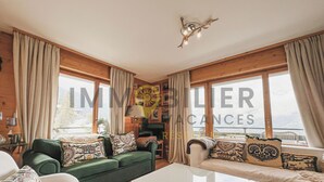 A spacious living room with a green sofa, a white coffee table, and a beige sofa. There are large windows with curtains, a fireplace, and a ceiling light fixture. - Situé à l'entrée du village, à proximité immédiate du bus navette et des commerces, ce lumineux et s (St-Luc)