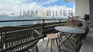 A balcony with wicker furniture, a table, and a view of a mountainous landscape. - Situé à l'entrée du village, à proximité immédiate du bus navette et des commerces, ce lumineux et s (St-Luc)