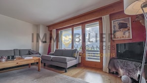 A living room with a sofa, a coffee table, and a TV. There is a window with a view of mountains.