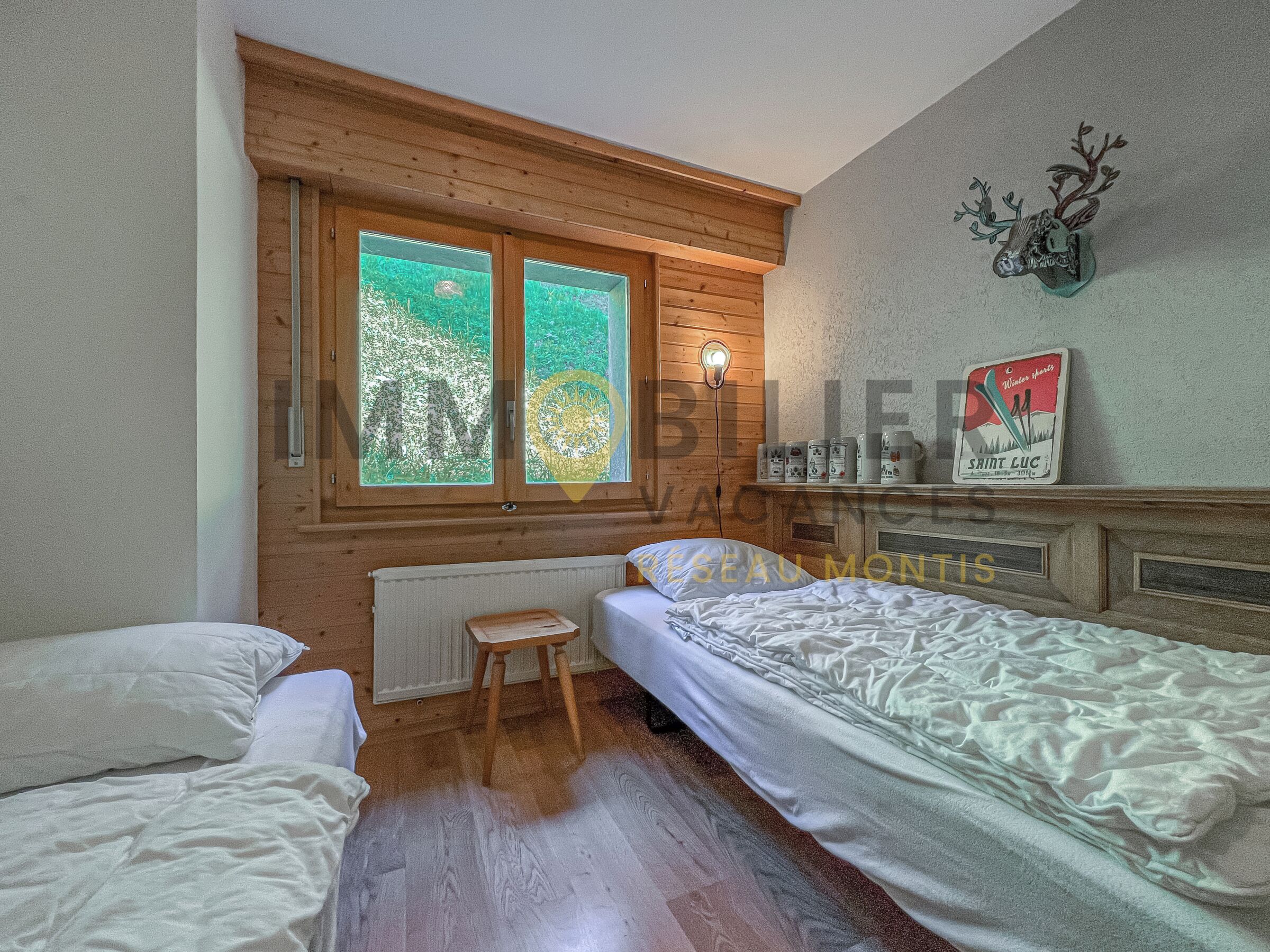 Ein gemütliches Zimmer mit zwei Betten, einem kleinen Hocker, einem Heizkörper und einem Regal mit Gegenständen.
