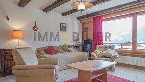 A cozy living room with a sofa, armchairs, a wooden coffee table, and a red rug. There is a lamp, a clock, and a window with a view of mountains. - Situé à l'entrée du village, à proximité immédiate du bus navette et des commerces. Idéal pour 2 à 4 (St-Luc)