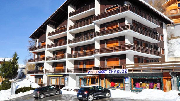 A multi-story hotel with balconies, a ski slope in the background, and a sign for 'INTERSPORT CHABLOZ'. - Situé à l'entrée du village, à proximité immédiate du bus navette et des commerces. Idéal pour 2 à 4 (St-Luc)