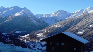 A wooden cabin with a snow-covered roof, surrounded by a mountainous landscape with snow-covered peaks. - Situé à l'entrée du village, à proximité immédiate du bus navette et des commerces. Idéal pour 2 à 4 (St-Luc)