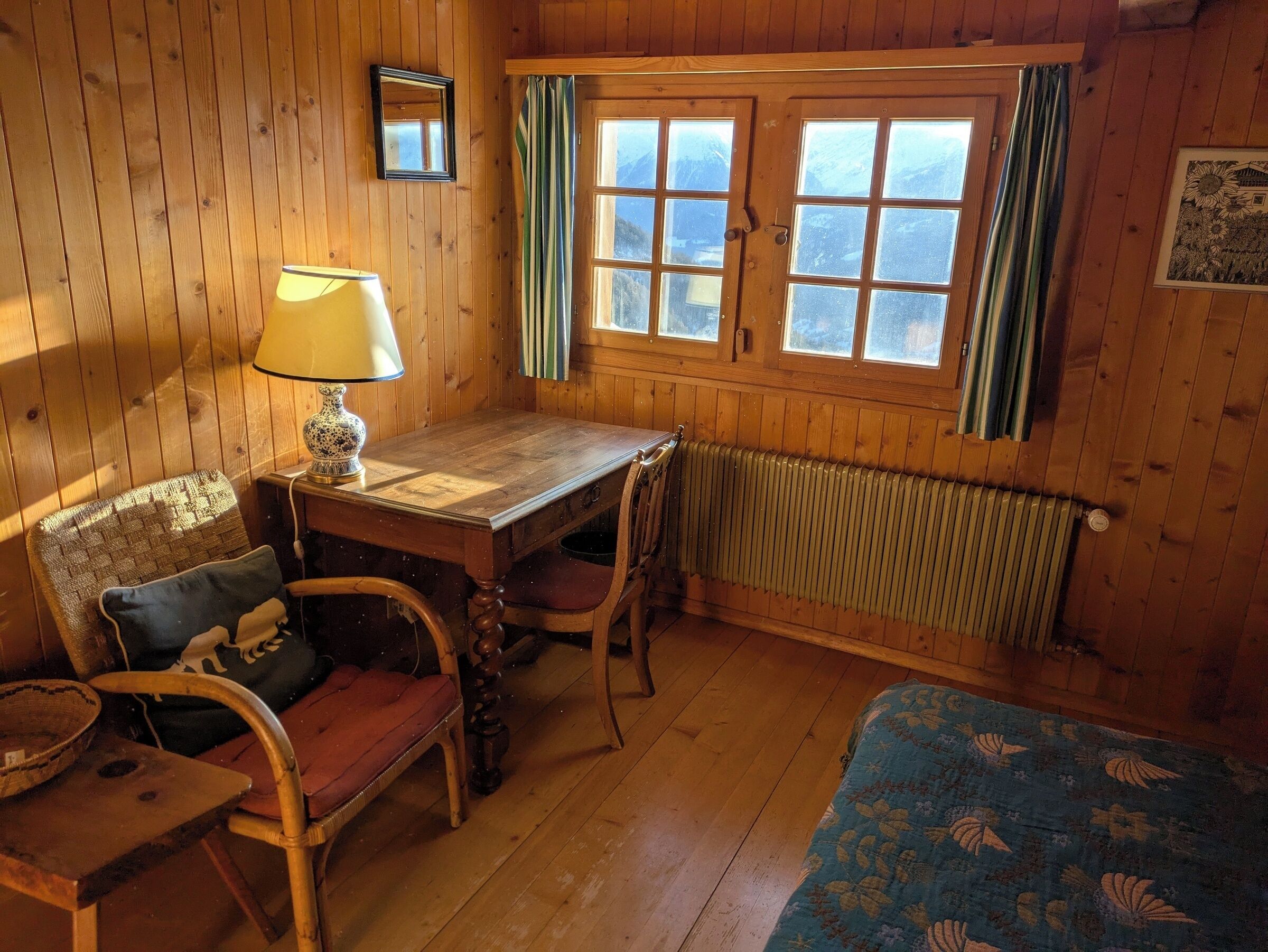 A cozy room with wooden walls and a large window, a table with a lamp, a chair, and a radiator.