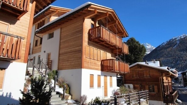 A wooden chalet with balconies, a stone staircase, and a snowy landscape. - Très beau chalet récent de tout confort situé directement au vieux village, avec un accès aisé. Idéa (St-Luc)