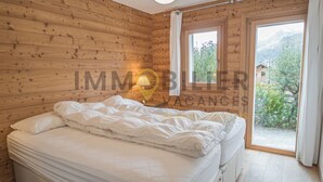 A wooden cabin bedroom with a bed, a nightstand, and a window offering a view of greenery and mountains. - Très beau chalet récent de tout confort situé directement au vieux village, avec un accès aisé. Idéa (St-Luc)
