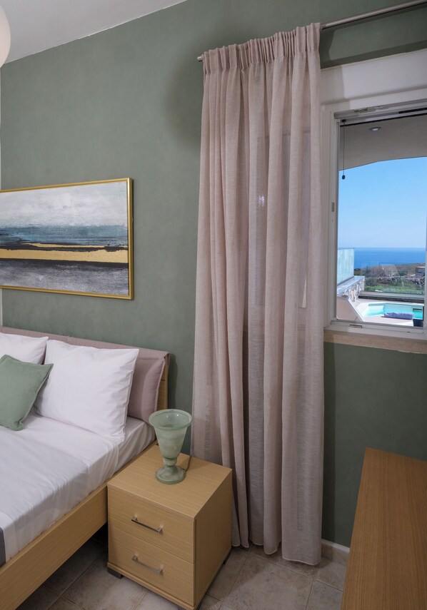 A bedroom with a bed, a bedside table, a nightstand, a painting, and a window with a view of the sea.