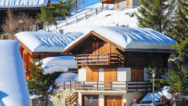 A wooden chalet with a balcony, surrounded by snow-covered trees and a clear sky.