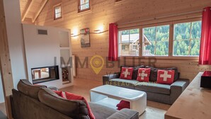 A cozy cabin-style room with a fireplace, a sofa set, and a large window offering a view of the forest. - Magnifique chalet confortable, moderne, de 195m2, 5 pièces  avec 4 chambres et pouvant accueillir ju (St-Luc)