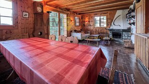 A wooden cabin interior with a dining table, sofa, fireplace, and large windows.