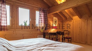 Un dormitorio tipo cabaña de madera con una cama, un escritorio y una silla. Hay ventanas con cortinas, un ventilador de techo y un cuadro en la pared.