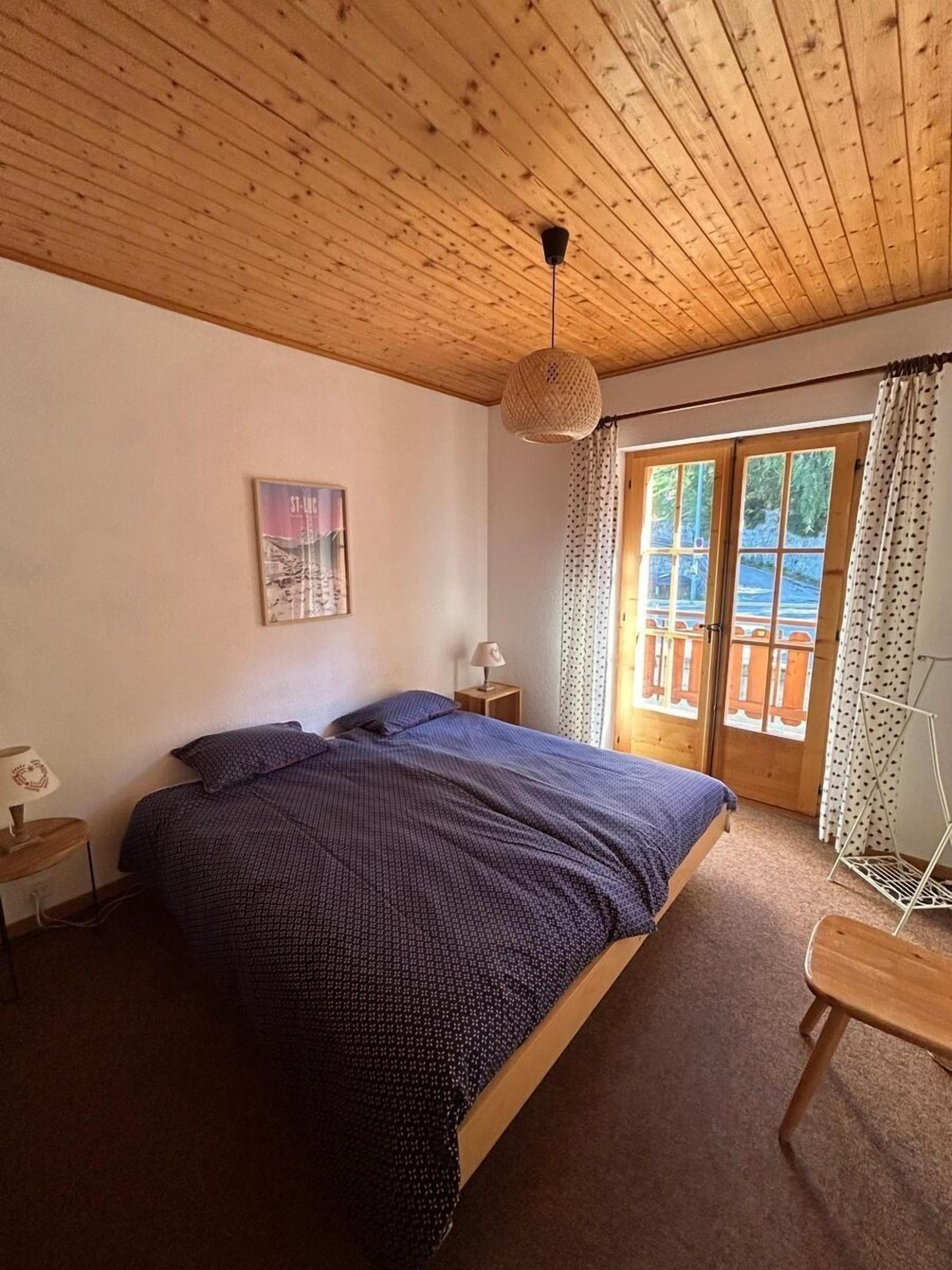 Une chambre avec un plafond en bois, un lit recouvert d’une courtepointe bleue, une table de chevet avec une lampe, un tableau accroché au mur et une fenêtre avec des rideaux.