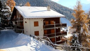 A chalet-style building with a sloped roof, wooden balconies, and a snow-covered landscape. - Idéalement situé, au pied des pistes, en face du funiculaire et à 5' à pied du centre du vieux villa (St-Luc)