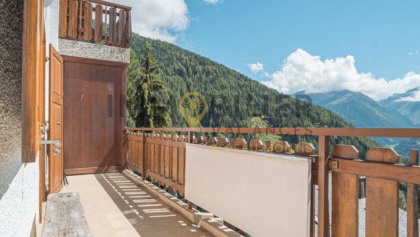 A balcony with a wooden railing, a wooden door, and a view of a forested mountain range. - Idéalement situé, au pied des pistes, face au funiculaire et à 5 minutes à pied du centre du vieux v (St-Luc)