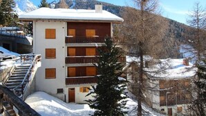 A multi-story building with wooden balconies, a snow-covered roof, and a mountainous backdrop. - Idéalement situé, au pied des pistes, face au funiculaire et à 5 minutes à pied du centre du vieux v (St-Luc)