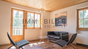 A room with a wooden ceiling, a black leather sofa, two foldable chairs, a small glass table, and large windows with a view of trees. - Idéalement situé, au pied des pistes, face au funiculaire et à 5 minutes à pied du centre du vieux v (St-Luc)