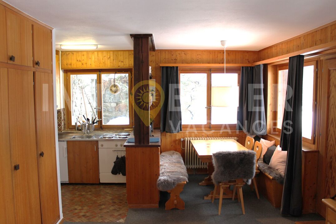 En stuga med träväggar, ett kök med spis och diskho, en matplats med en bänk och stora fönster med gardiner.