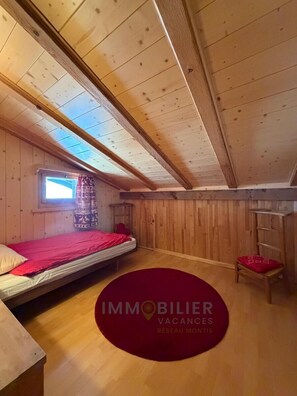 A small, wooden room with a single bed, a window, and a red circular rug.