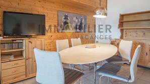A wooden-paneled room with a round dining table, chairs, a television, and a bookshelf. - Duplex de 3,5 pièces (75 m2) pour 4-5 personnes très lumineux, situé en face de l’Office du tourisme (Chandolin)