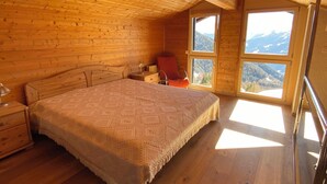 A cozy wooden cabin bedroom with a large bed, a nightstand, a chair, and a view of mountains through the windows. - Duplex de 3,5 pièces (75 m2) pour 4-5 personnes très lumineux, situé en face de l’Office du tourisme (Chandolin)