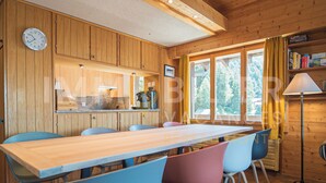 A wooden cabin interior with a dining table, chairs, a clock, and a window with curtains. - Situé au centre de Chandolin dans une petite résidence, proche de toutes commodités et du bus navett (Chandolin)