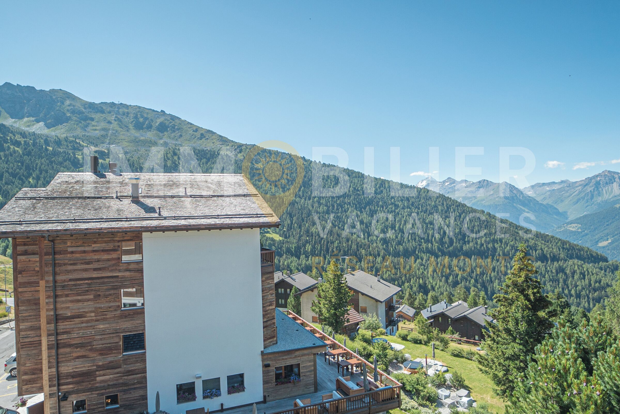 Un edificio moderno con fachada de madera y un balcón, rodeado de un paisaje montañoso.