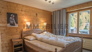 A wooden-paneled bedroom with a bed, bedside lamps, a nightstand, and a window with curtains. - Situé au centre de Chandolin dans une petite résidence, proche de toutes commodités et du bus navett (Chandolin)