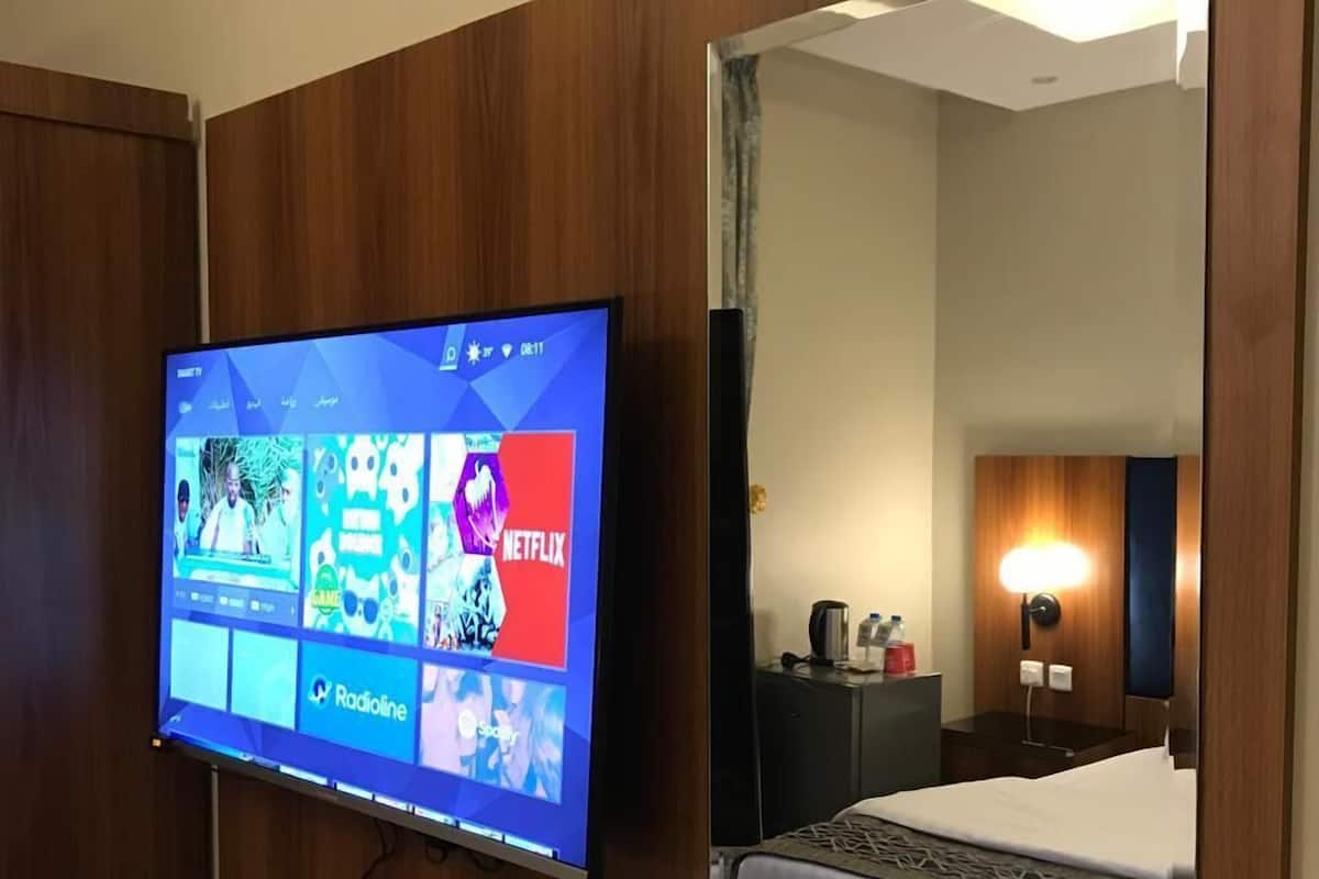 A hotel room with a flat-screen TV mounted on the wall, a bed with a white comforter, a bedside table with a lamp, and a mirror reflecting the room.