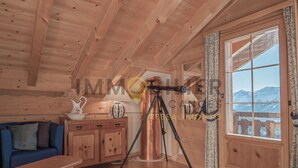 Interior de una cabaña de madera con un telescopio, un sofá azul, armarios de madera y una ventana con vistas a la montaña.