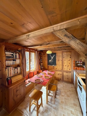 A wooden cabin interior with a dining table, chairs, bookshelves, and a kitchen area.