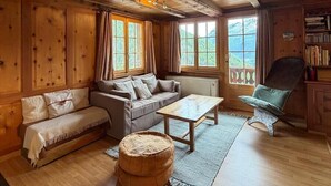 A cozy cabin interior with a wooden ceiling, a sofa, a table, a chair, and a bookshelf.