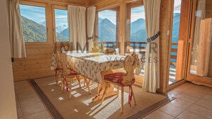 Una cabaña de madera con comedor que incluye mesa y sillas, ventanas grandes con cortinas y vistas a las montañas.