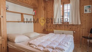 A wooden-paneled room with a bed, a chair, and a window with curtains. - Spacious 2.5-room apartment with TV and Wi-Fi, south-west facing, with 2 large balconies, ideal for (St-Luc)