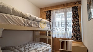 A bunk bed room with a wooden headboard, a window with patterned curtains, and a radiator. - Spacieux et lumineux 3.5 pièces pour 4 à 6 personnes, situé au 1er étage (angle ouest) d'une résiden (St-Luc)