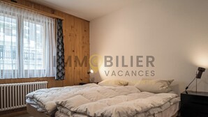 A bedroom with a wooden headboard, a bed with white bedding, a bedside table with a lamp, and a window with sheer curtains. - Spacieux et lumineux 3.5 pièces pour 4 à 6 personnes, situé au 1er étage (angle ouest) d'une résiden (St-Luc)