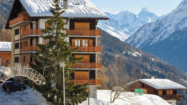 A multi-story chalet with wooden balconies, set against a backdrop of snow-covered mountains. - Spacieux et lumineux 3.5 pièces pour 4 à 6 personnes, situé au 1er étage (angle ouest) d'une résiden (St-Luc)