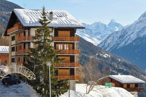 A multi-story chalet with wooden balconies, set against a backdrop of snow-covered mountains. - Spacieux et lumineux 3.5 pièces pour 4 à 6 personnes, situé au 1er étage (angle ouest) d'une résiden (St-Luc)