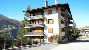 A multi-story building with balconies, a parking area, and a mountainous backdrop. - Spacieux et lumineux 3.5 pièces pour 4 à 6 personnes, situé au 1er étage (angle ouest) d'une résiden (St-Luc)