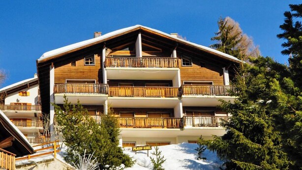 A multi-story wooden building with balconies, likely a hotel or lodge, surrounded by snow and trees.