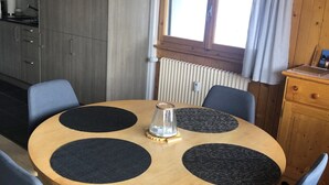 A round wooden table with four black placemats, surrounded by six blue chairs. A small glass vase is on the table.