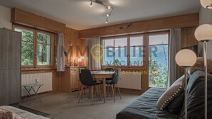 A cozy room with a wooden interior, a dining table with chairs, a sofa, and large windows offering a view of greenery.