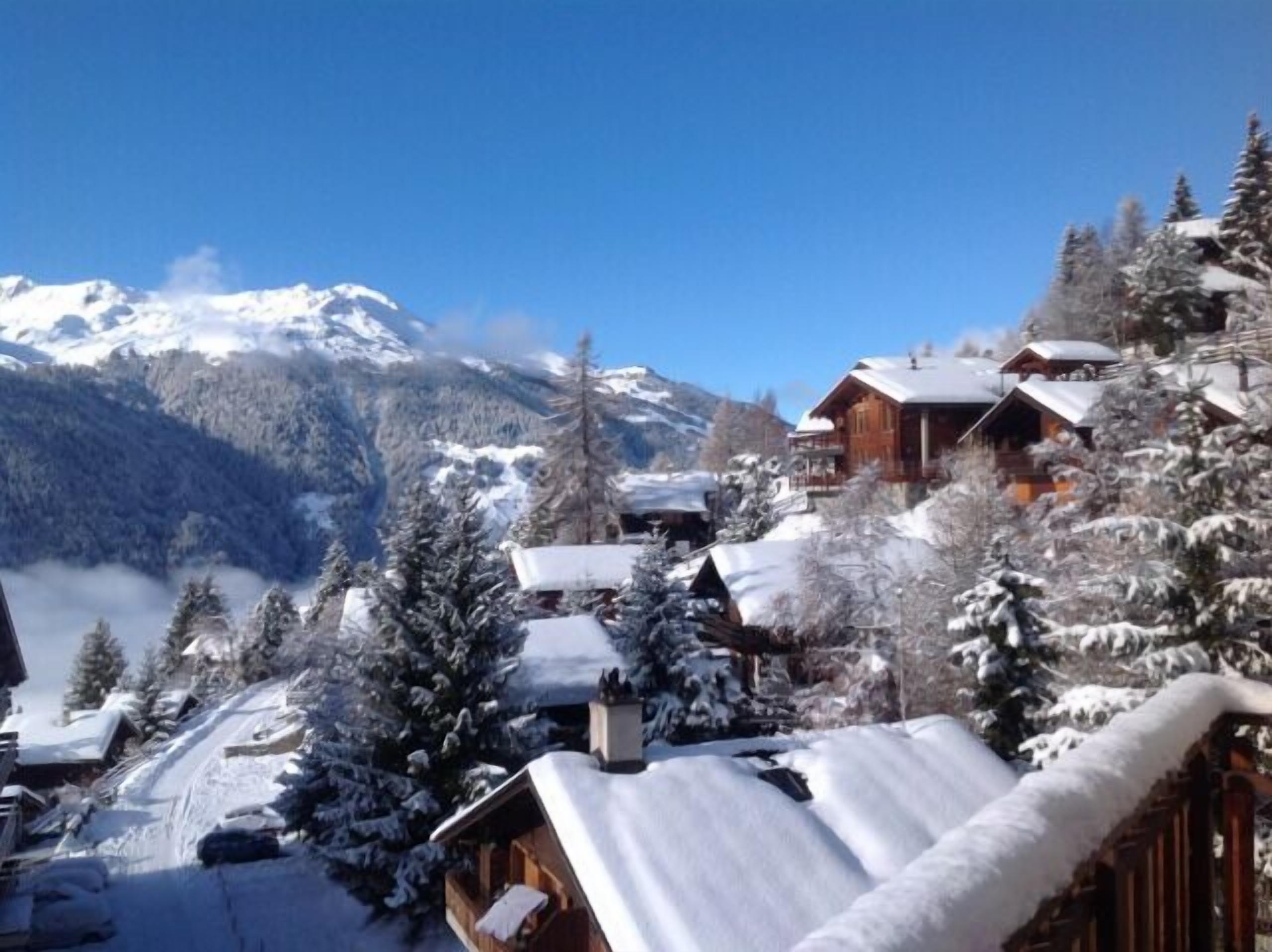 A snow-covered mountain village with wooden cabins and a clear view of snow-capped peaks in the background.