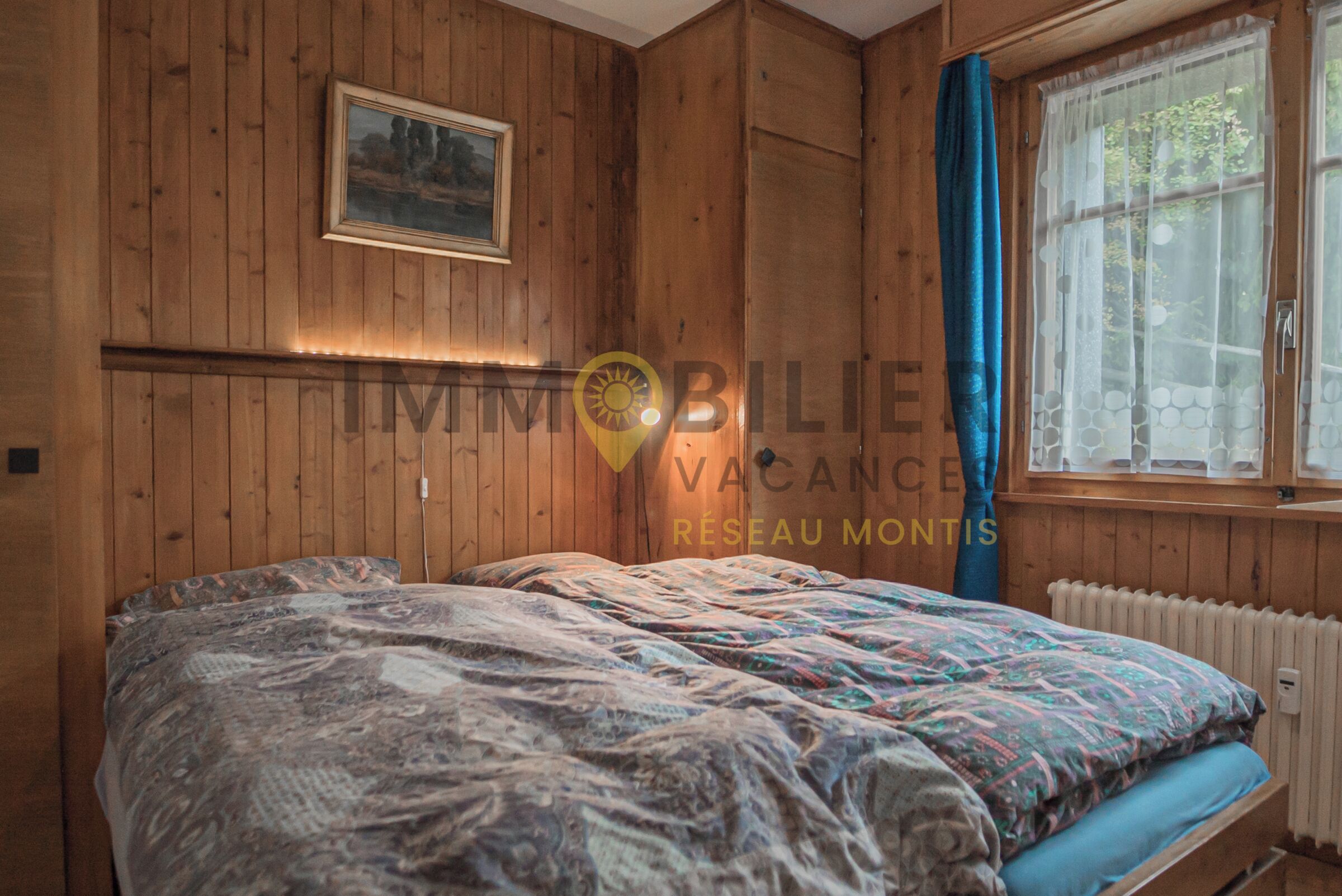Ein Zimmer mit Holzwänden, einem Bett, einem Fenster mit Vorhängen und einer Heizung.