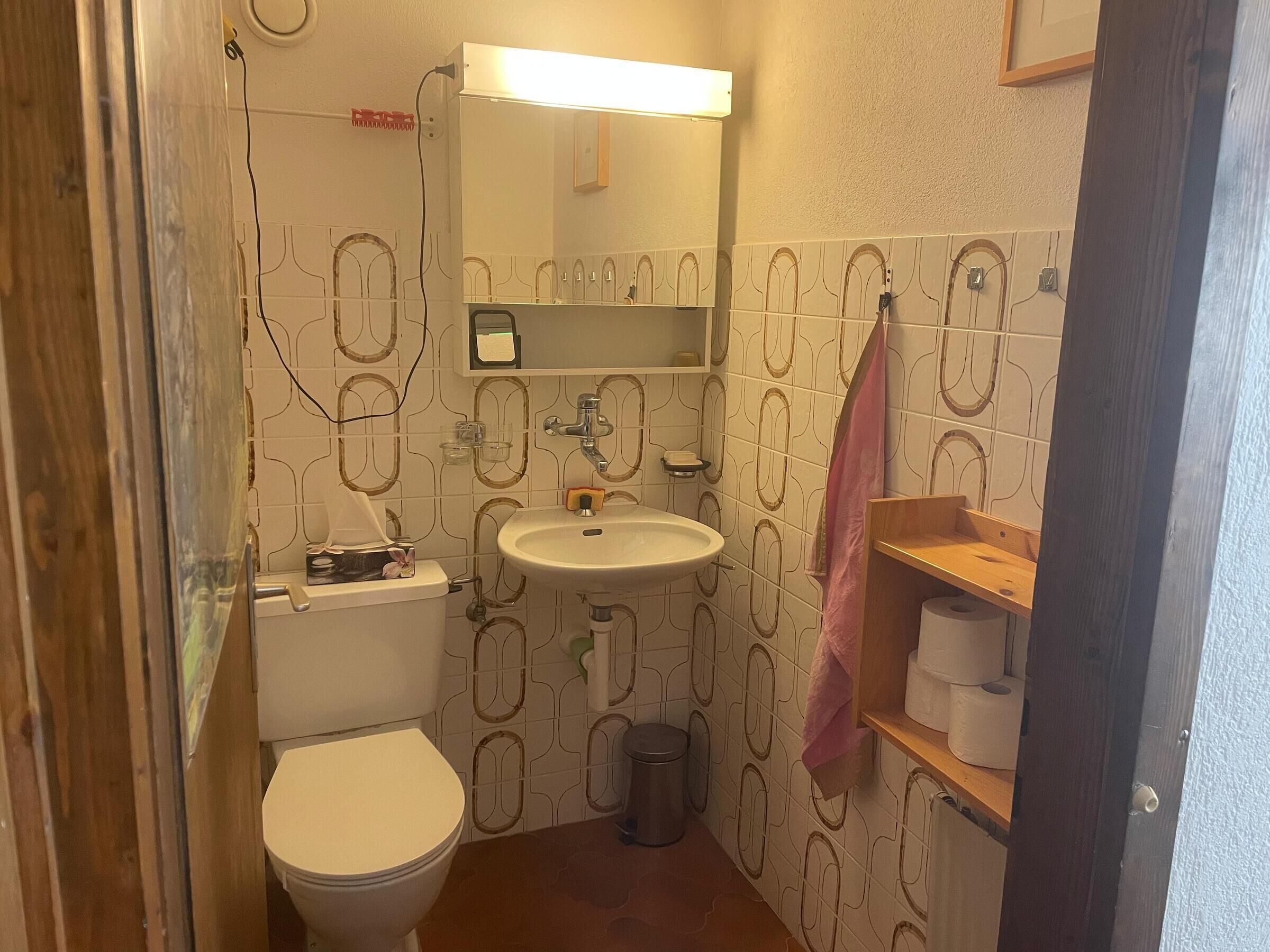 A small bathroom with a toilet, sink, and shower. There is a towel hanging on the wall and a trash can.