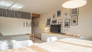 A modern living room with a sofa, a dining table, and a kitchen area. There are framed pictures on the wall and a ceiling light fixture.