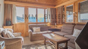 A cozy living room with wooden walls and large windows, furnished with sofas and a coffee table.