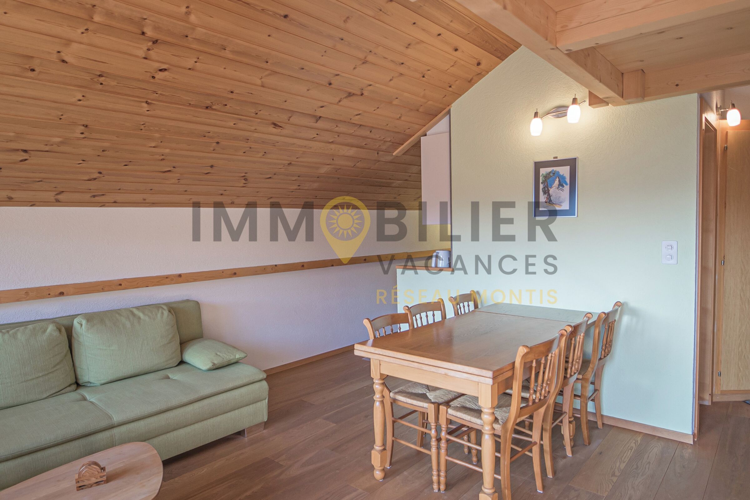 Una habitación con techo de madera, un sofá verde, una mesa de comedor de madera con sillas, y una pared de color claro con un cuadro enmarcado.