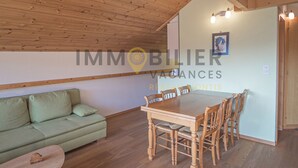 Ein Zimmer mit Holzbalkendecke, einem grünen Sofa, einem Holzesstisch mit Stühlen und einer hellen Wand mit einem gerahmten Bild.